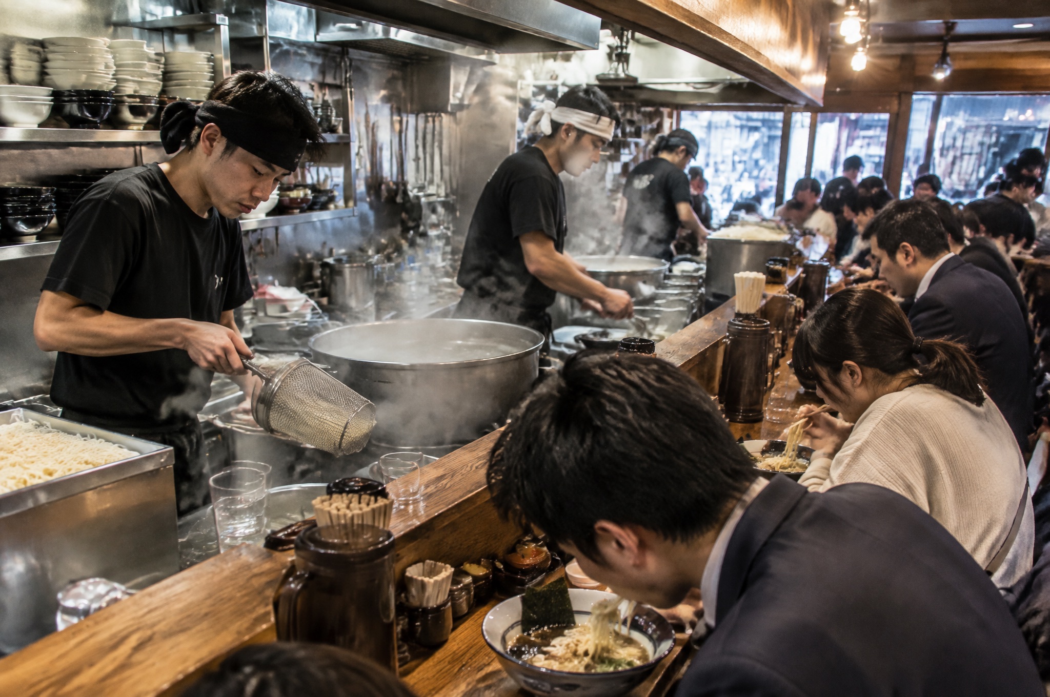 昼のピーク時に湯気の中で働くラーメン店のスタッフ。AI生成のイメージで、実際の画面・事例・提供内容を示すものではありません。